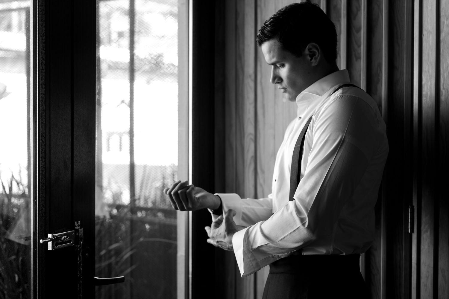 Black and white photo of a well-dressed groom adjusting his cufflinks by a window in natural light.