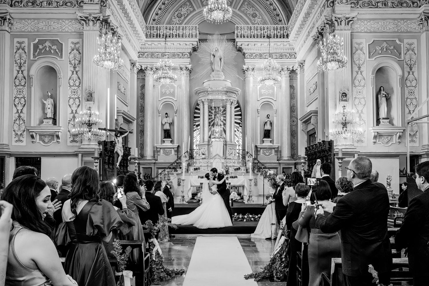 Black and white photo of a wedding ceremony inside an ornate church with guests seated as couple stands at the altar.