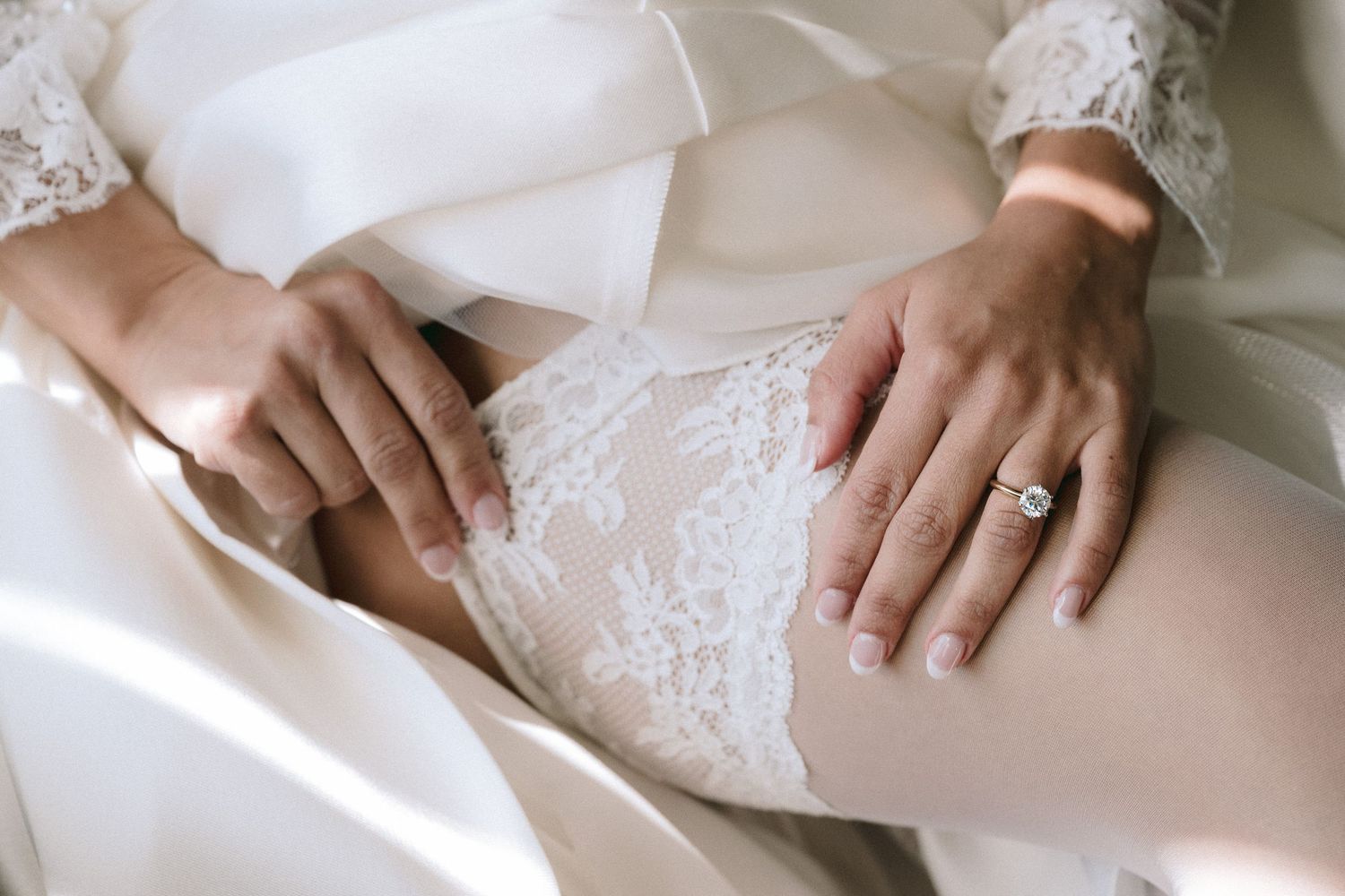 Hands resting on white lace wedding dress showing elegant engagement ring.