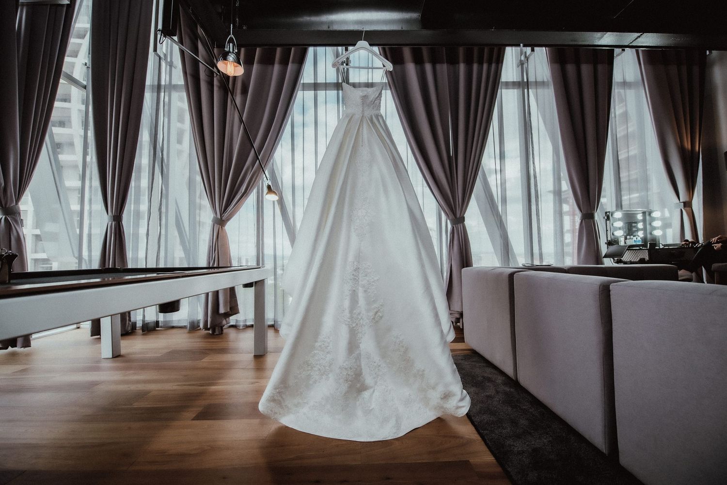 Elegant white wedding dress displayed in a room with gray curtains and natural lighting.