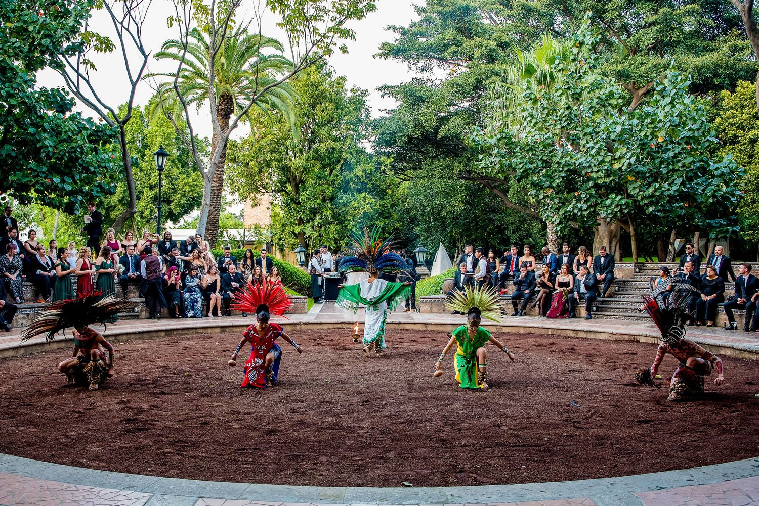Traditional Aztec dancers in colorful headdresses perform in a garden plaza before an elegant crowd of spectators.