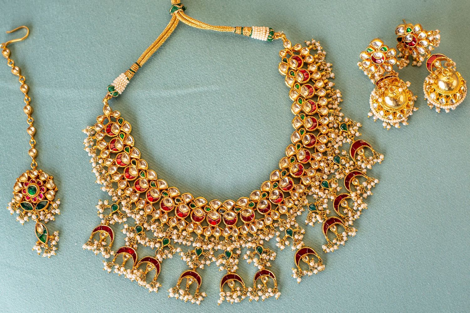 Traditional Indian gold kundan bridal jewelry set with red gems, pearl dangles, jhumka earrings, and maang tikka on teal background.
