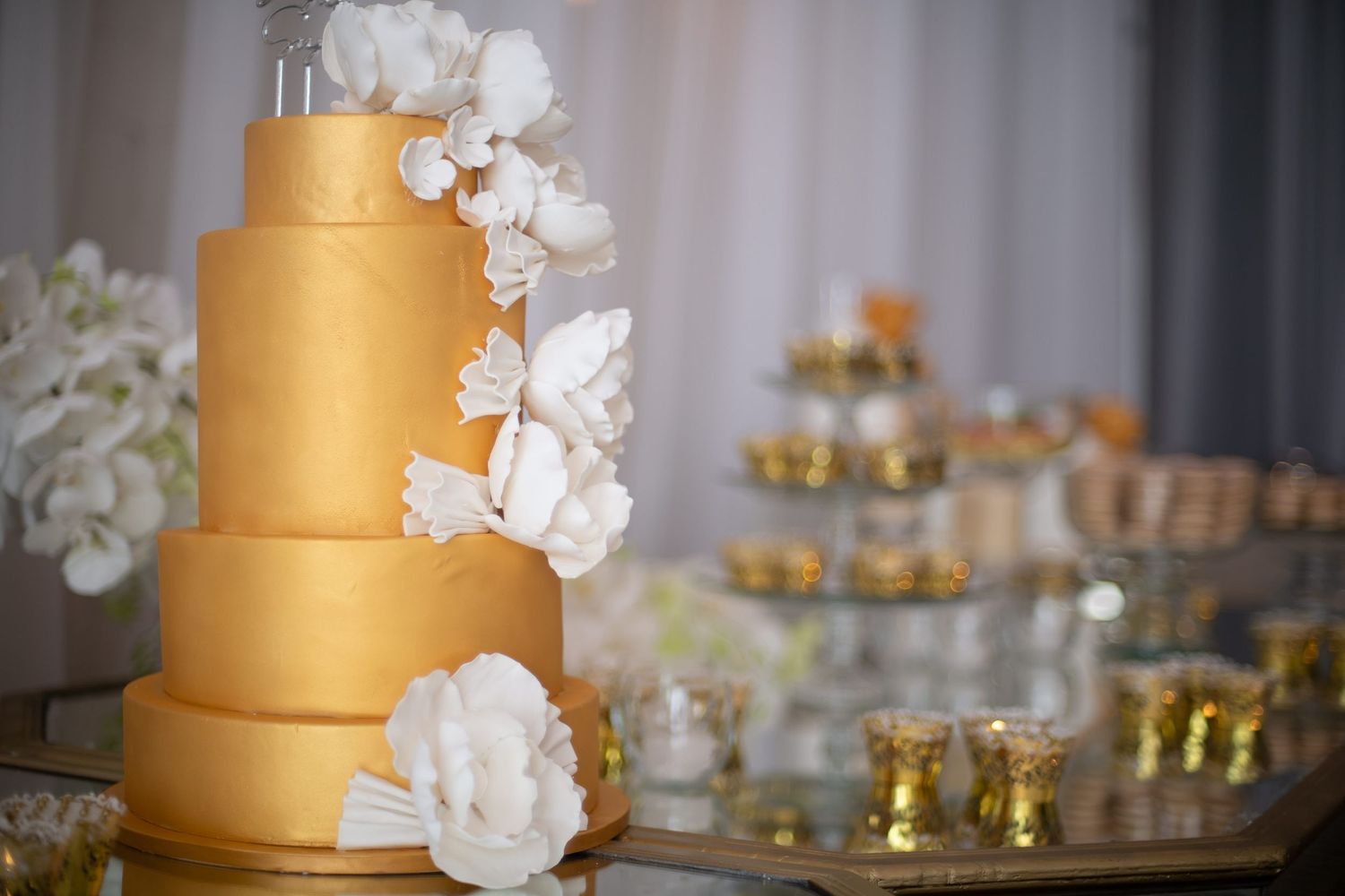 Elegant four-tier gold wedding cake adorned with white sugar flowers on a mirrored table with gold votive candles.