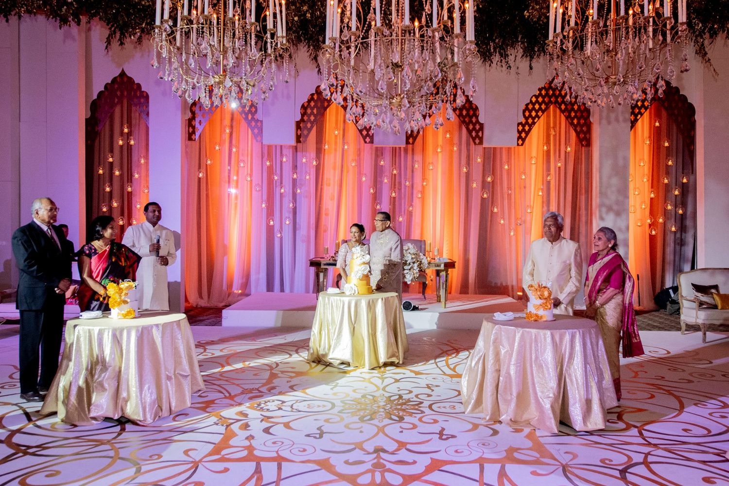 Elegant Indian wedding ceremony with chandeliers, floral drapes, and guests in traditional attire on ornate flooring.