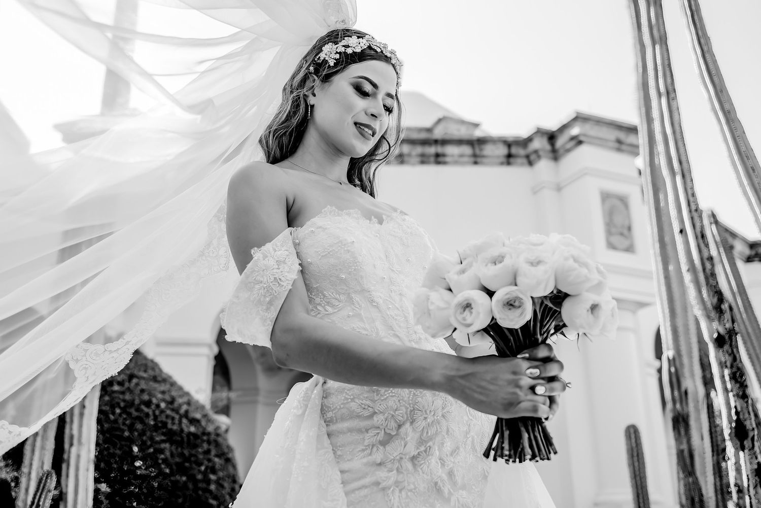 Black and white photo of a bride in a lace gown holding a white bouquet, with her veil flowing in the wind.
