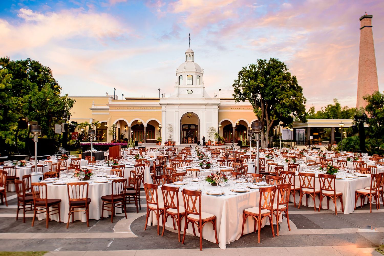 Elegant outdoor wedding reception with round tables, wooden chairs, and a white colonial venue backdrop at sunset.