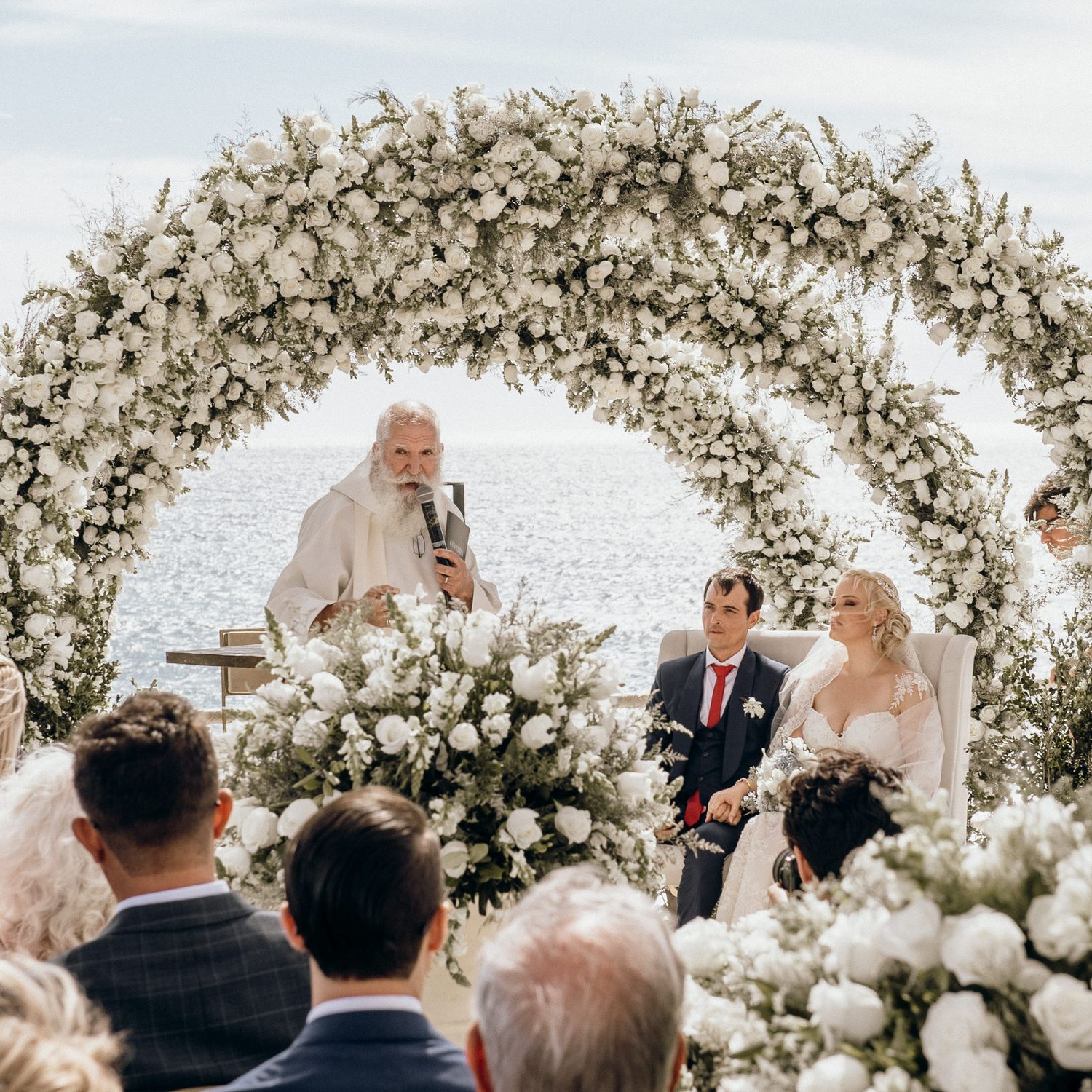 Outdoor wedding ceremony under an elegant white floral arch overlooking the ocean.