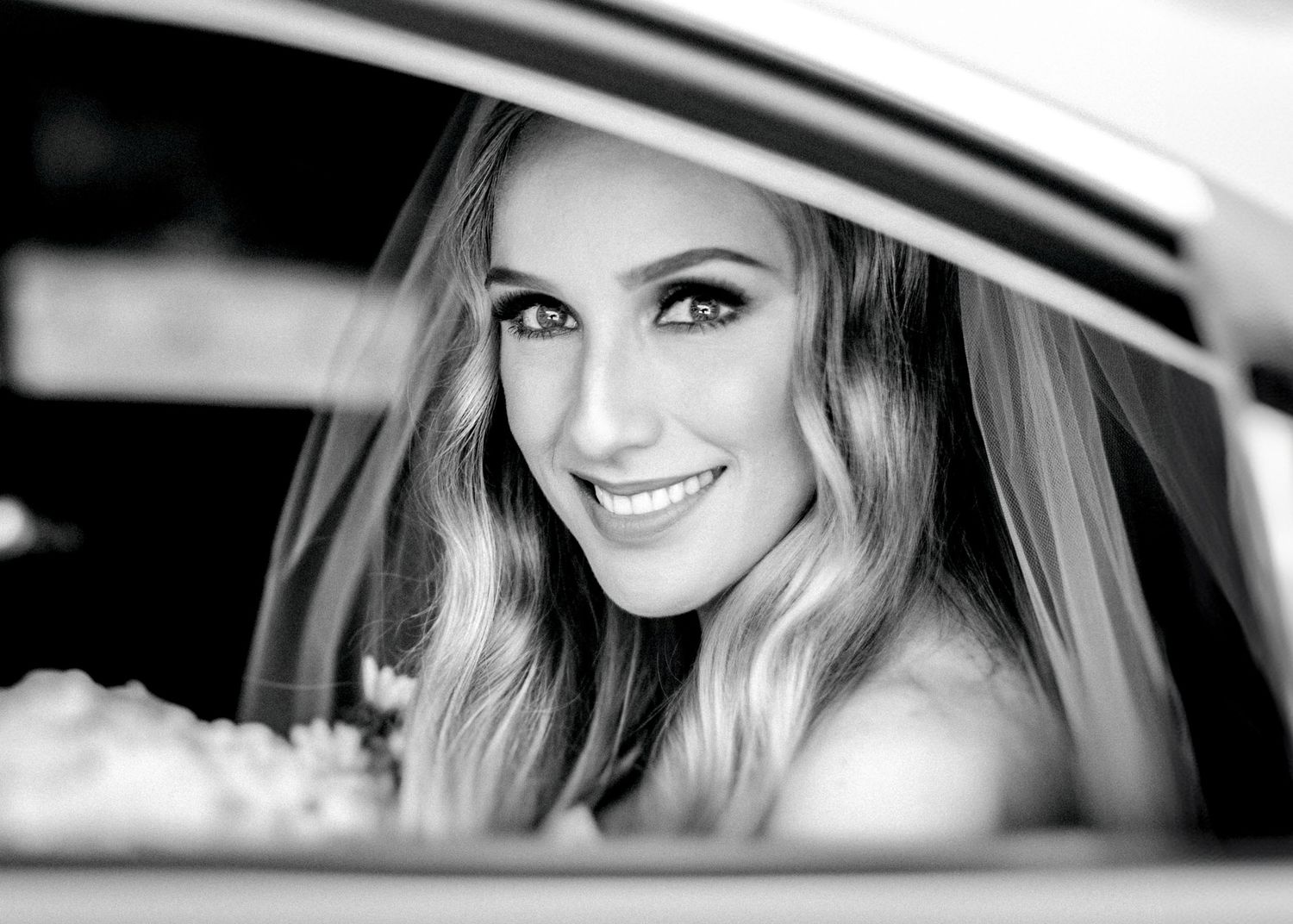 Black and white close-up of a bride with veil smiling through a car window.