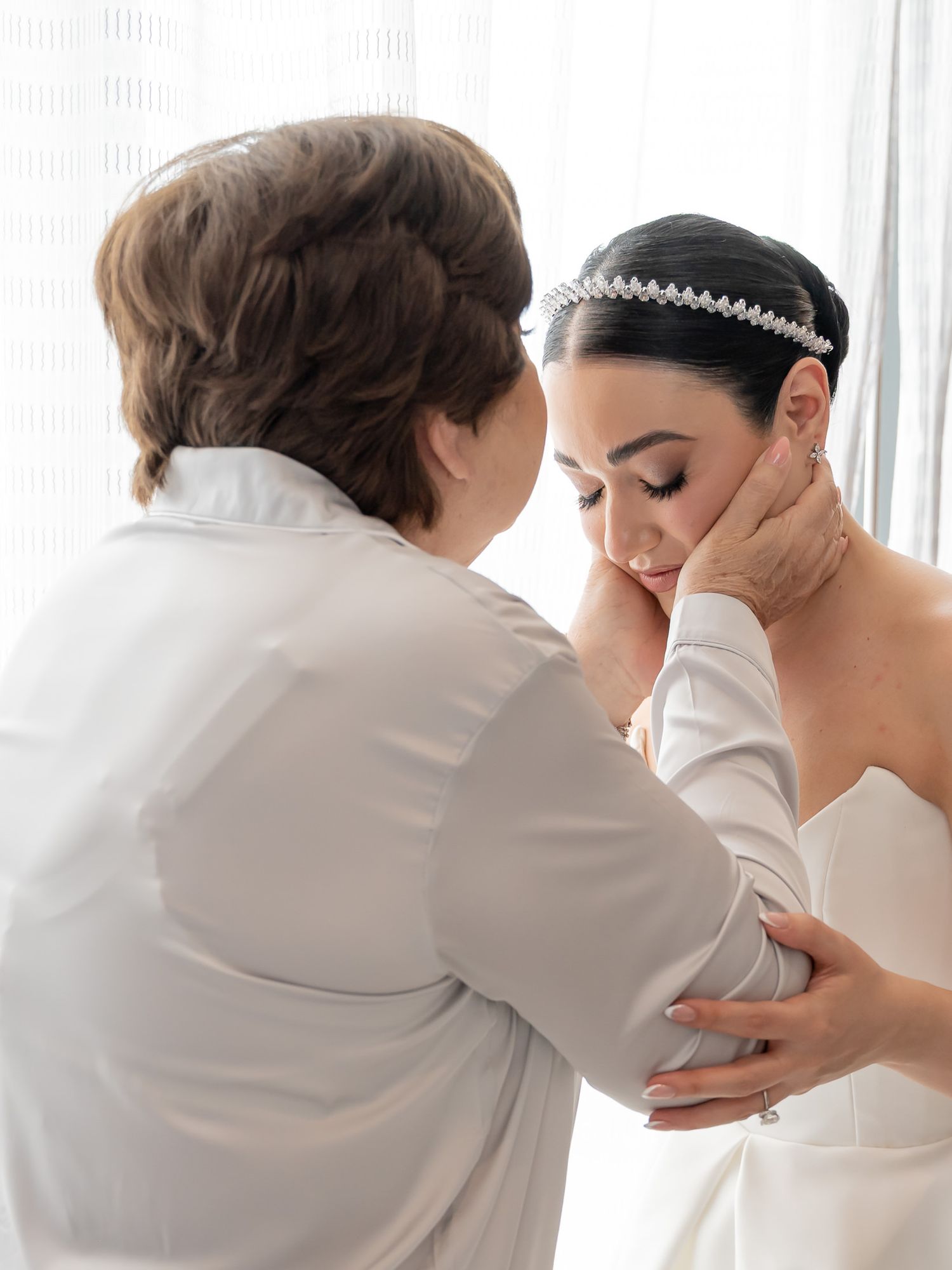 A bride in a strapless gown and crystal headband shares an emotional moment with an older woman in white.