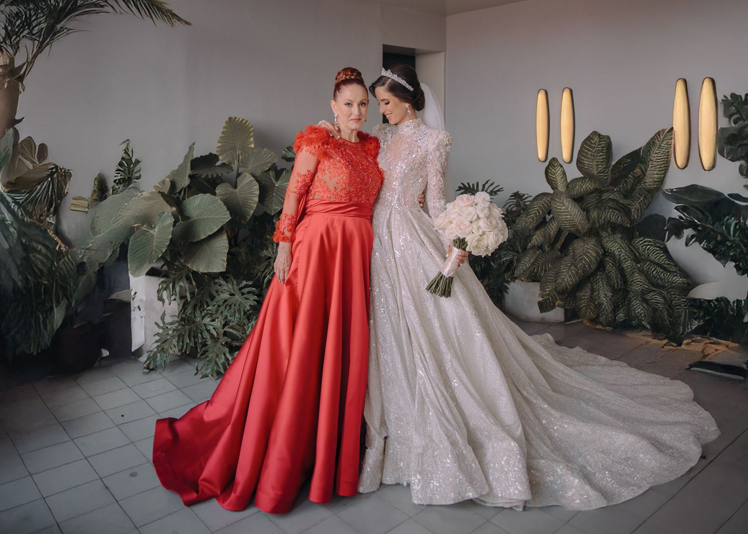 Two women pose together, one in a red ball gown and one in a beaded white wedding dress, surrounded by tropical plants.