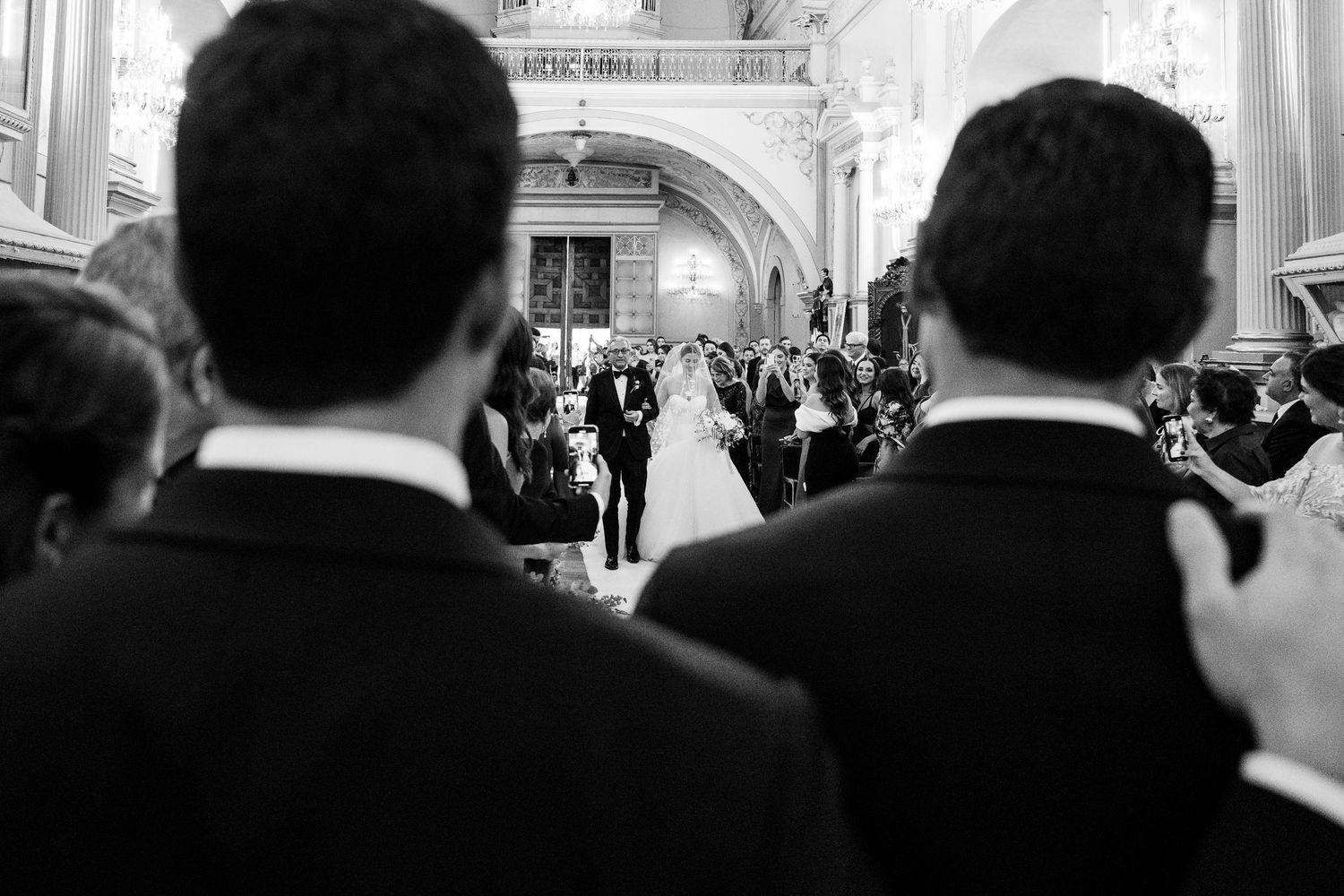 Black and white wedding ceremony photo showing bride and groom at altar with guests and groomsmen in foreground.