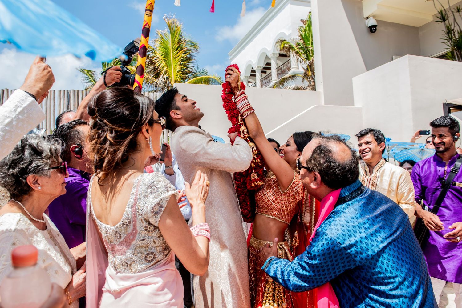 Joyful Indian wedding celebration with guests lifting the groom outdoors under a bright blue sky.