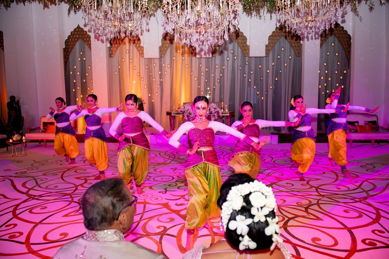 Traditional Indian dancers in colorful costumes perform on a pink-lit stage at an elegant cultural celebration.