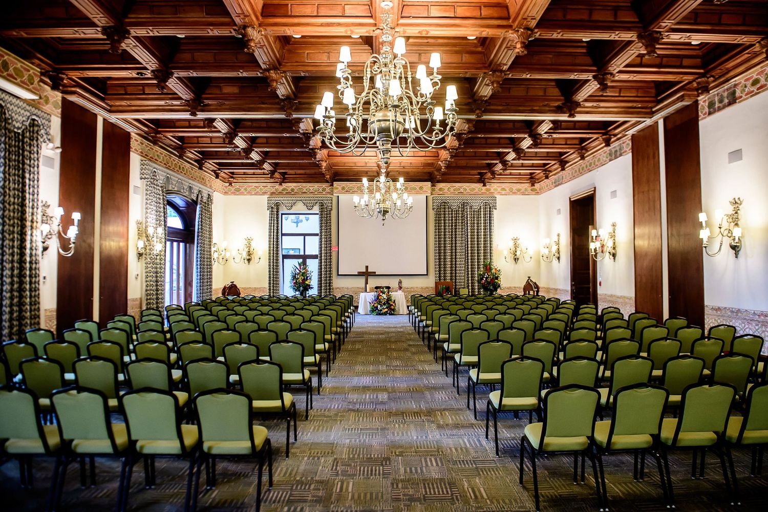 Elegant banquet hall with green chairs, ornate chandelier, wood ceiling, and floral altar setup for a ceremony.