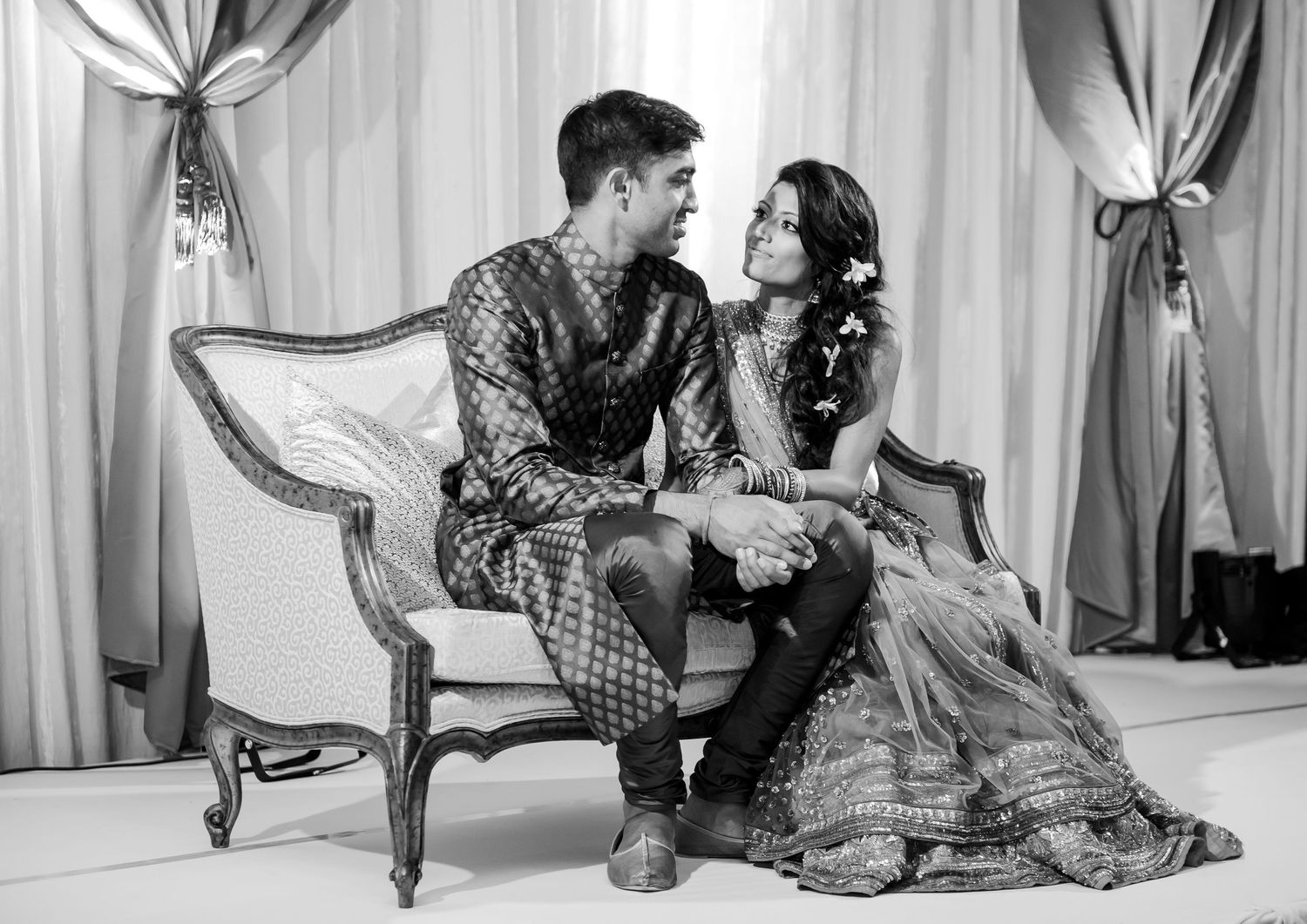 Black and white photo of an Indian couple in traditional attire sitting on an ornate sofa during a wedding ceremony.
