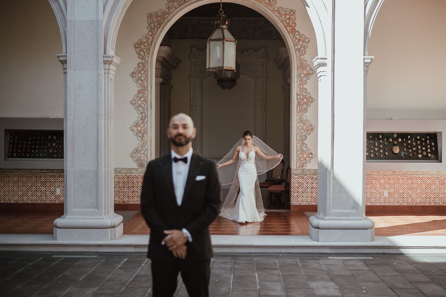 Groom in black tuxedo stands in foreground as bride in white gown and veil poses under arched colonnade.