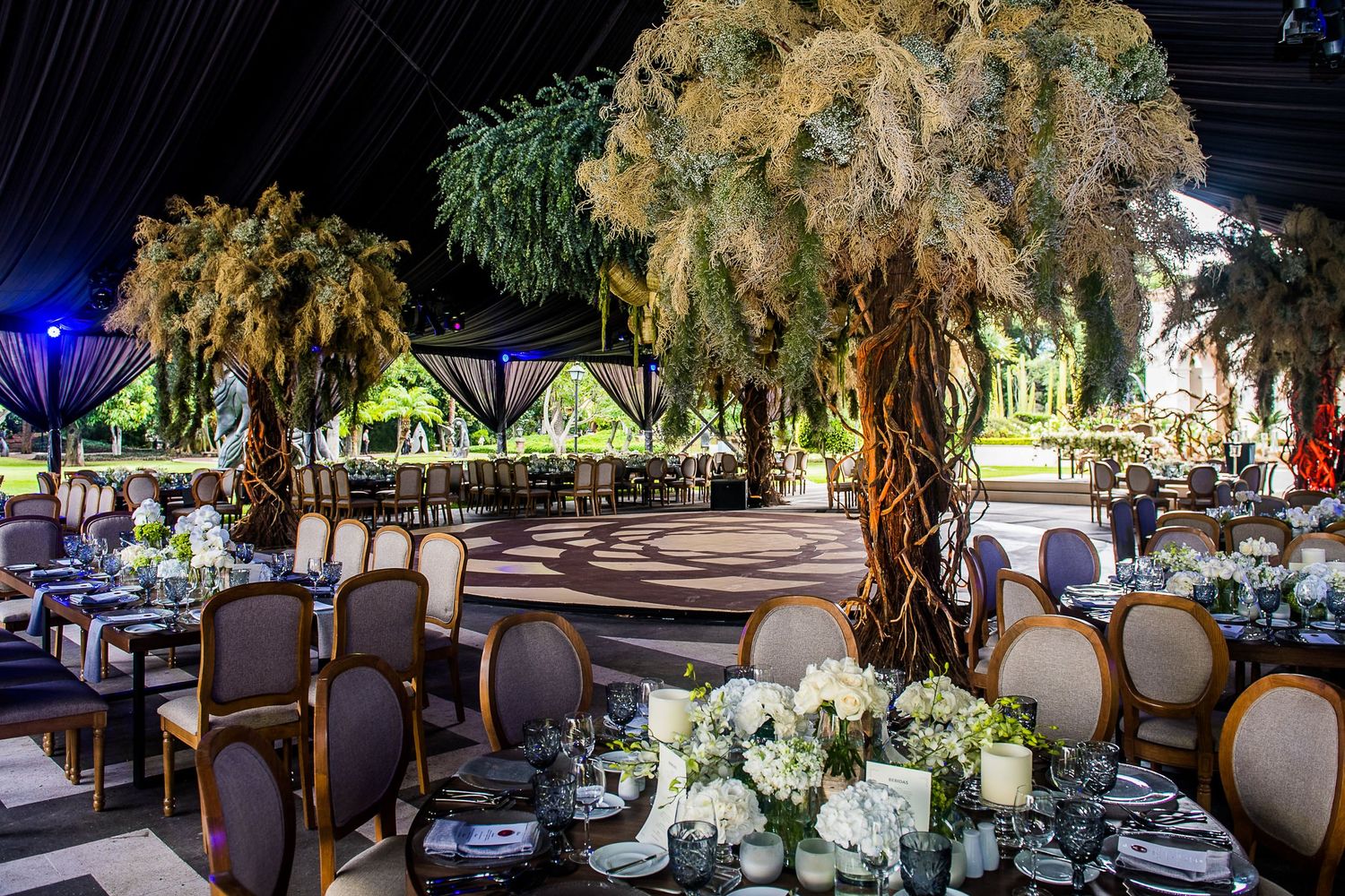 Elegant outdoor wedding reception with tall dried floral arrangements, round tables, white flowers, and candles under a dark tent.