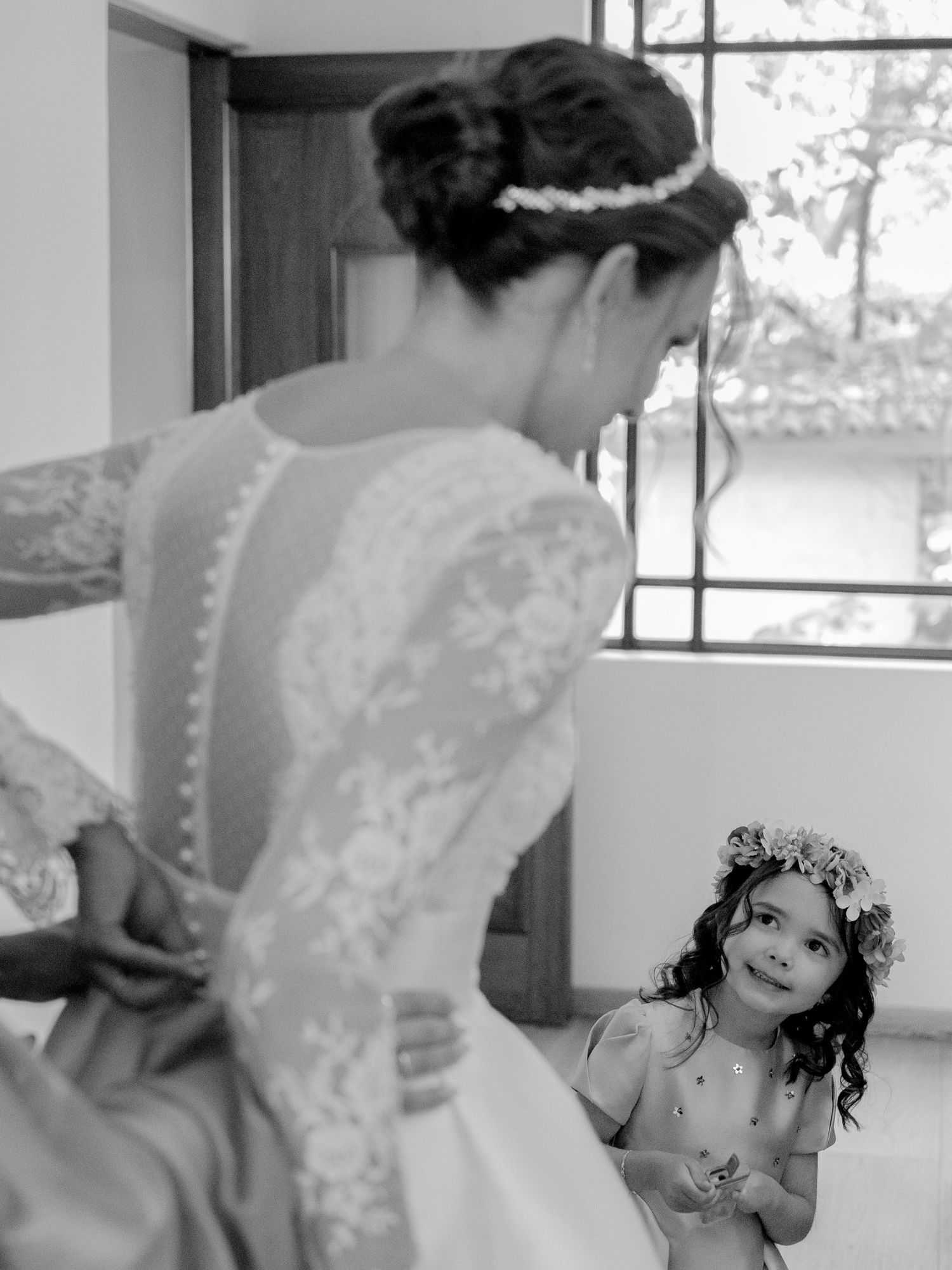 Black and white photo of a bride in lace gown with a flower girl in floral crown gazing up at her admiringly.