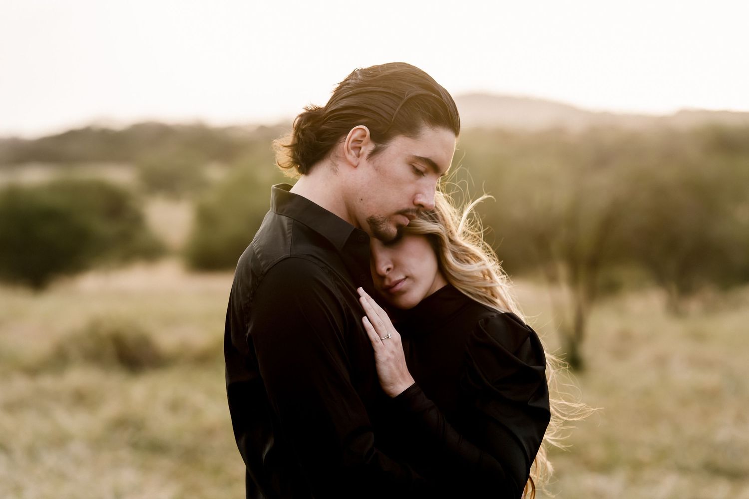 Couple embracing tenderly in a golden field, man in black kissing woman's forehead during romantic outdoor portrait.