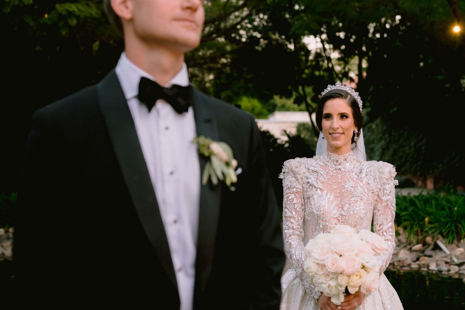 Bride in lace long-sleeve gown with tiara holds white bouquet, groom in black tuxedo with bow tie at outdoor wedding.