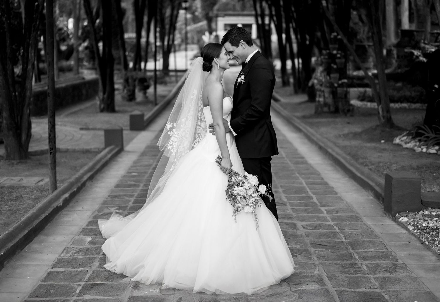 Black and white wedding photo of bride and groom sharing a romantic kiss on a tree-lined pathway.