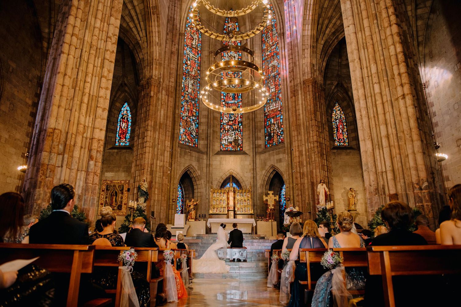 Wedding ceremony inside a grand Gothic cathedral with ornate chandeliers, stained glass windows, and guests seated in wooden pews.
