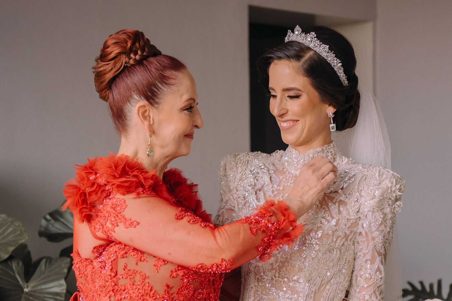 A bride in a beaded gown and tiara gets her necklace fastened by a woman in a red lace dress.