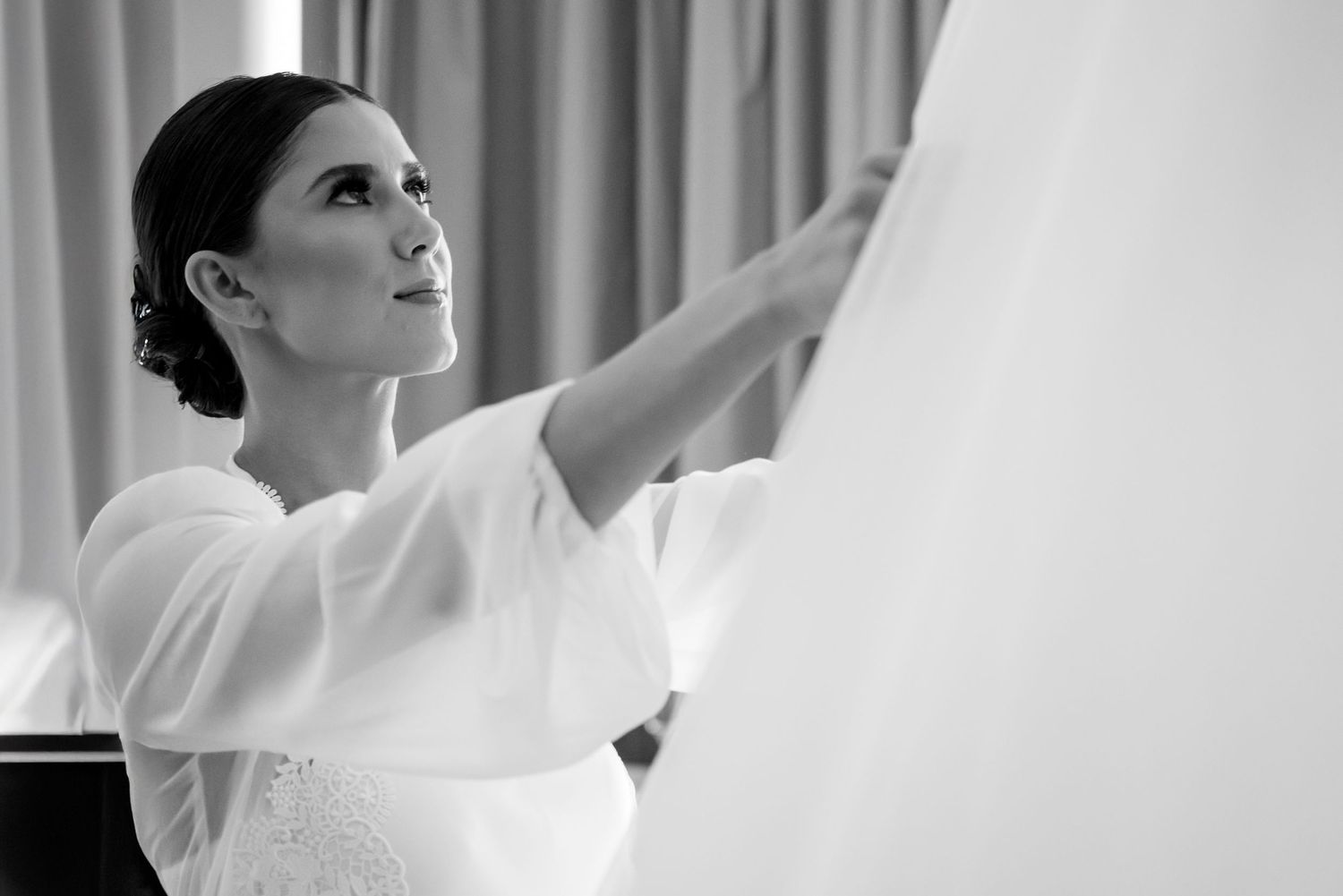 Black and white photo of a bride in white dress adjusting her wedding gown, elegant updo hairstyle.