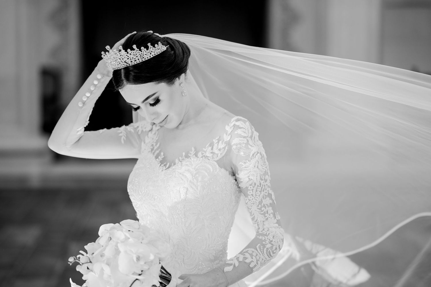 Black and white photo of a bride in lace gown and crown holding bouquet with veil flowing gracefully behind her.