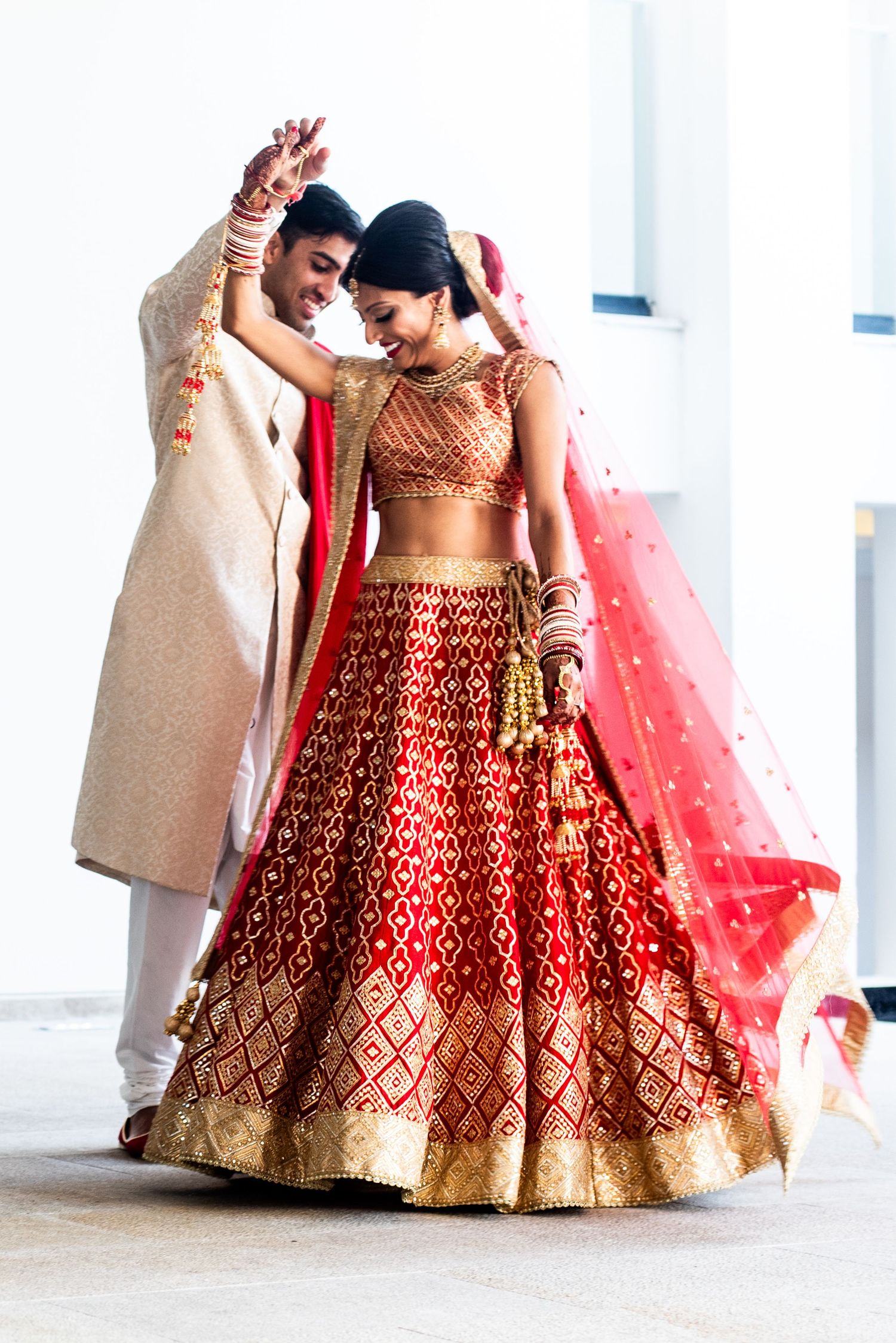 Indian bride in red and gold lehenga dances with groom in cream sherwani during traditional wedding ceremony.