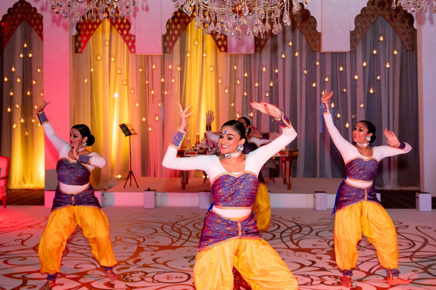 Three women perform a vibrant Indian classical dance in yellow and purple costumes at an elegant decorated event venue.
