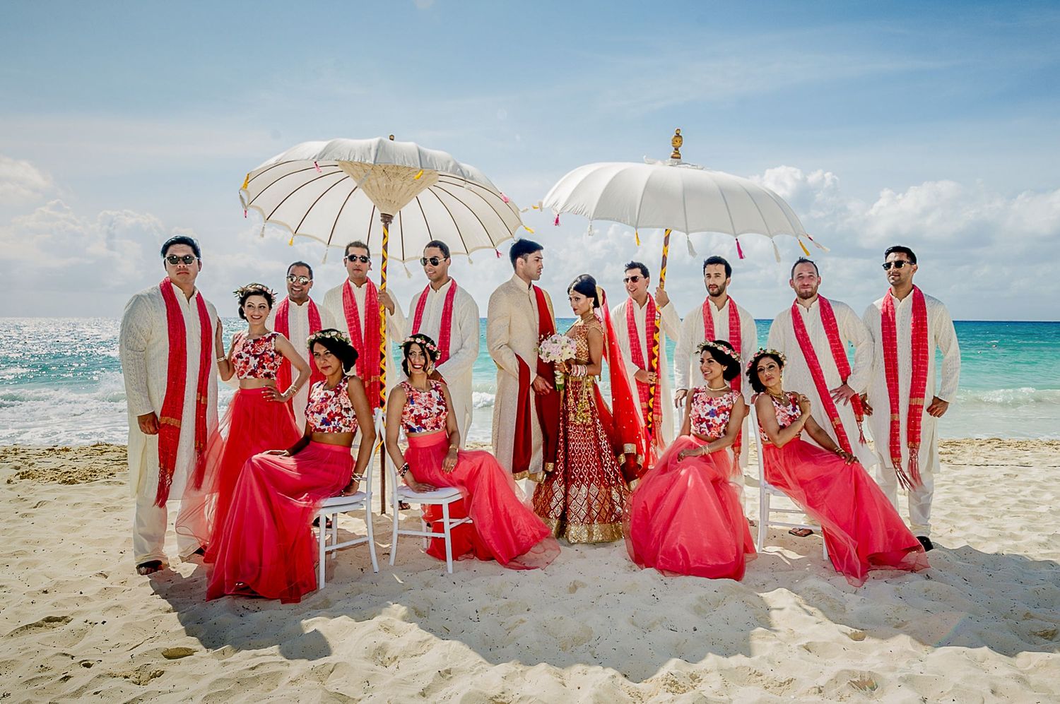 Beach wedding party with bridesmaids in red dresses posing with parasols on sandy shore.