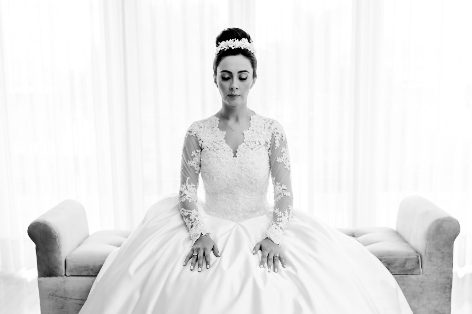Elegant black and white portrait of a bride in long-sleeved lace gown seated on white furniture.