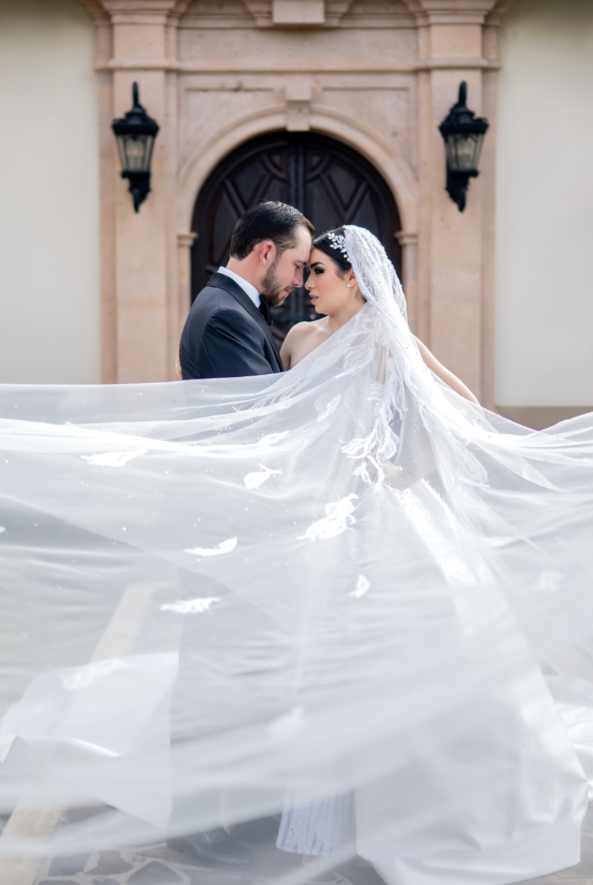 A wedding couple embraces as the bride's long white veil dramatically flows in the wind against a stone building backdrop.