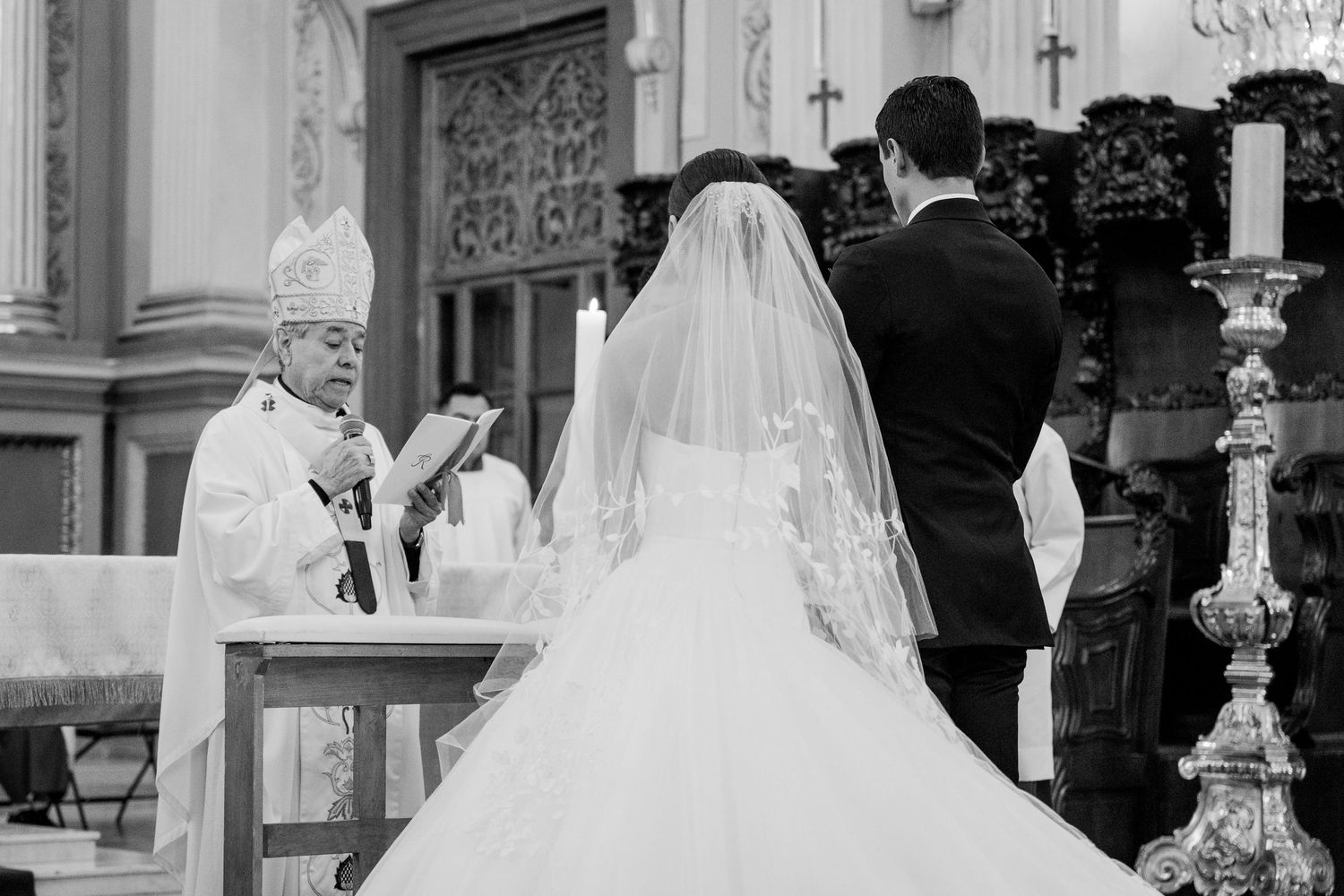 Black and white photo of a bride and groom exchanging vows before a bishop at a Catholic church wedding ceremony.