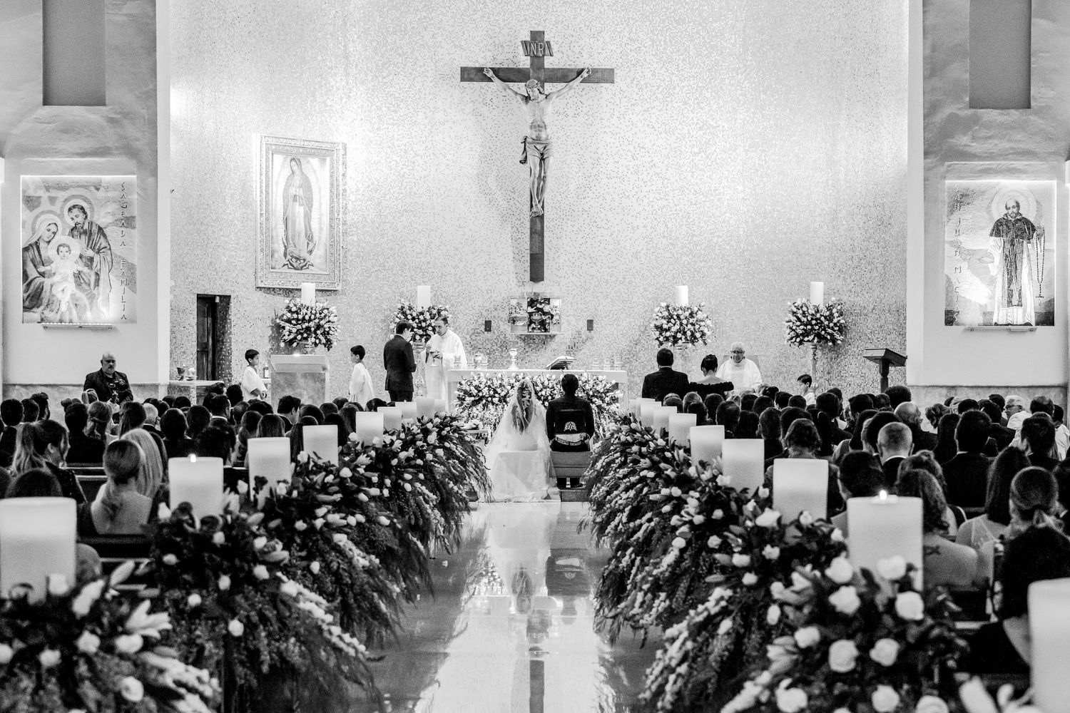 Black and white photo of a church wedding ceremony with floral aisle decorations and a crucifix altar.