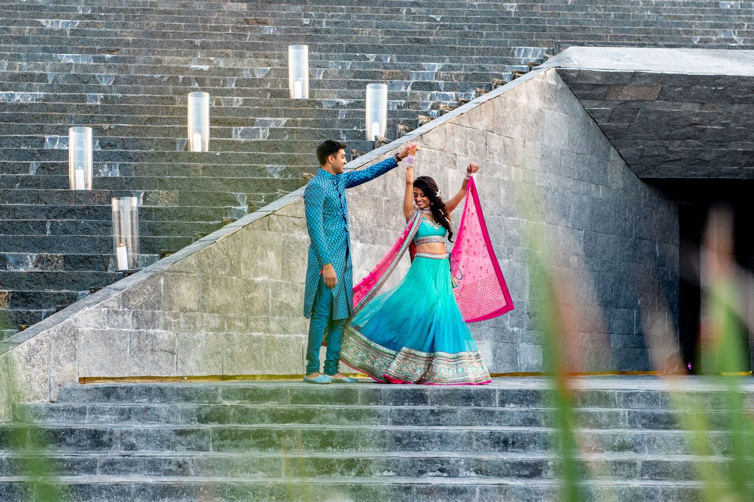 Couple in vibrant Indian wedding attire on stone steps, man in teal kurta twirling woman in blue and pink lehenga.