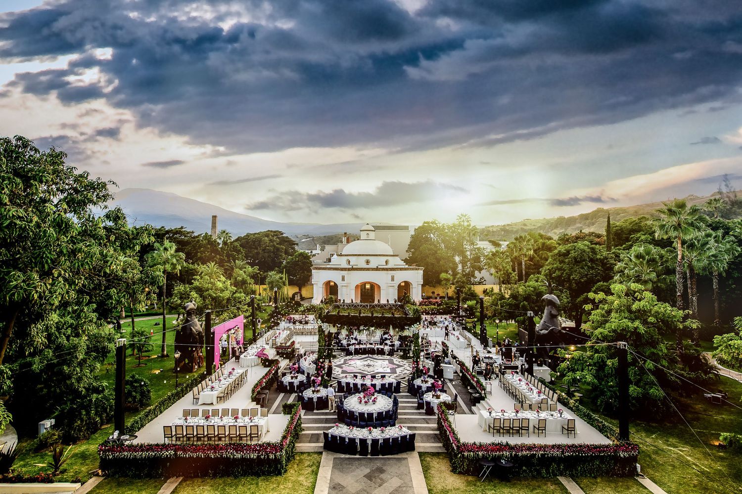 Aerial view of an elegant outdoor event venue with white tables, lush gardens, and a historic building at sunset.