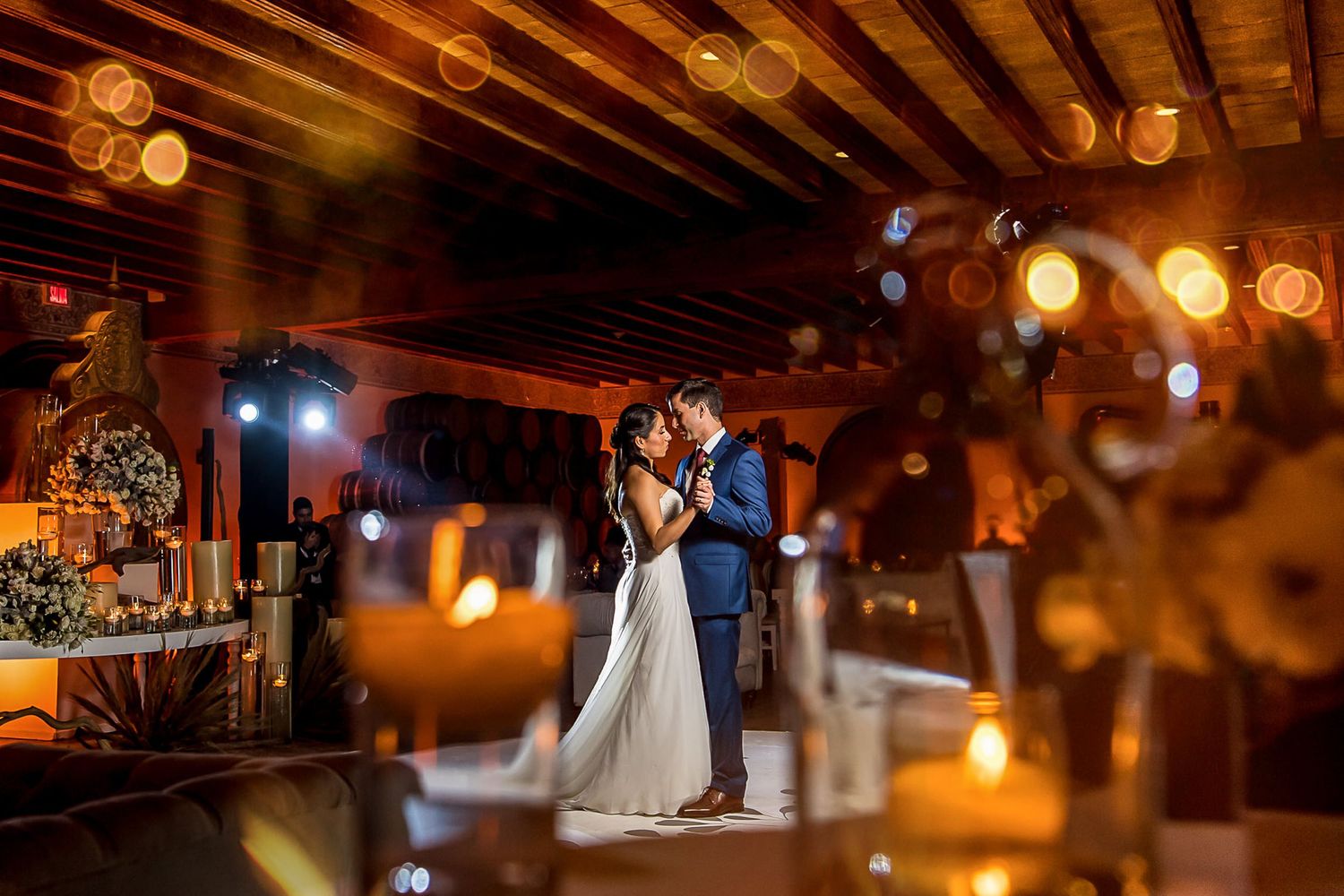 Romantic wedding couple sharing first dance in warmly lit rustic venue with candles and bokeh lights.