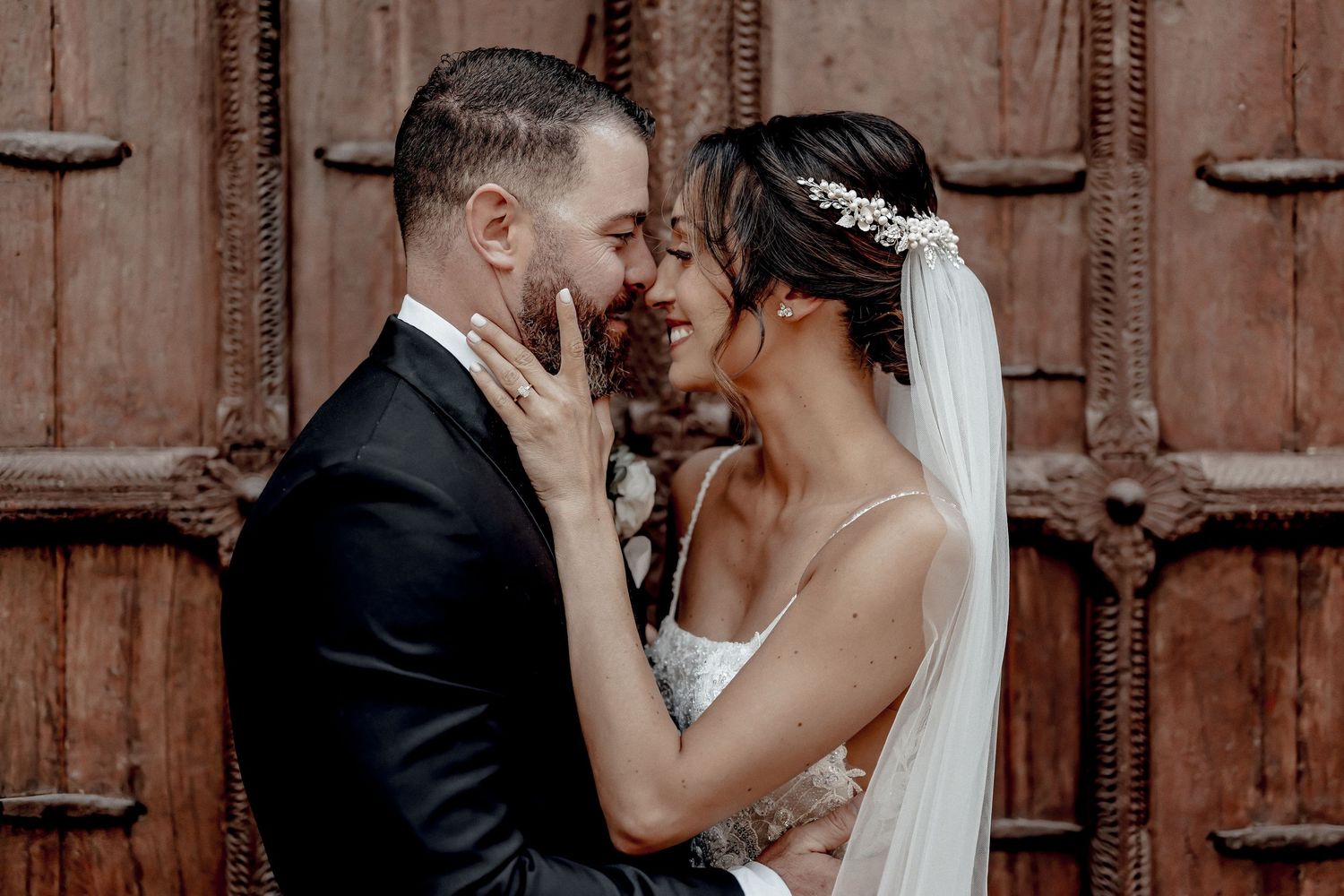 A bride and groom share a romantic moment, nearly kissing, in front of a rustic wooden door on their wedding day.