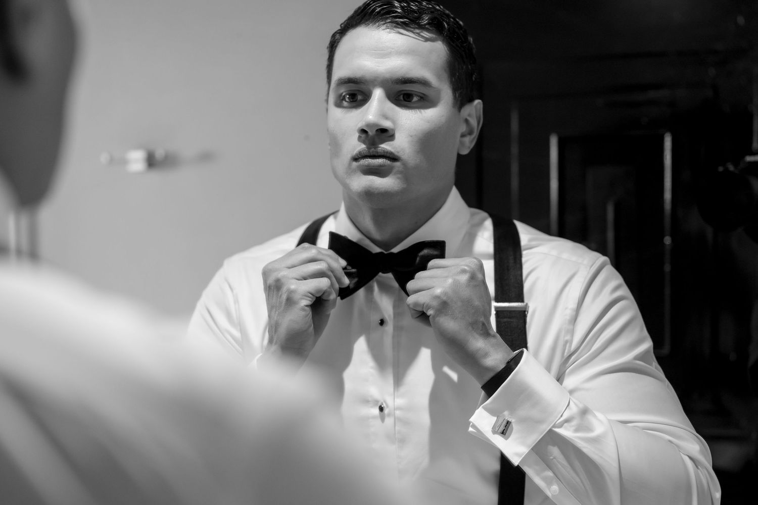 Black and white photo of a groom adjusting his bow tie while looking in a mirror, wearing suspenders and a dress shirt.
