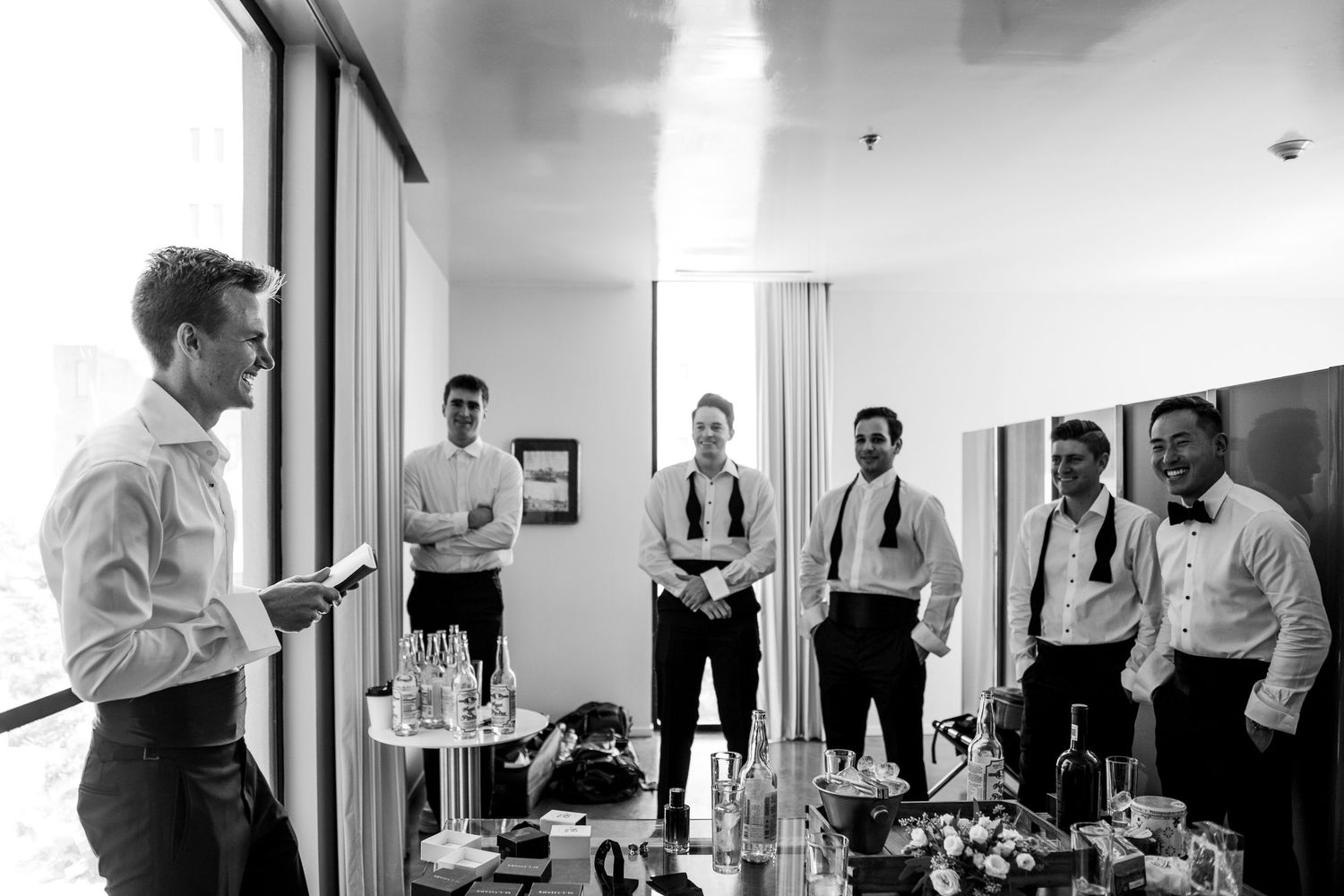Groomsmen in formal attire gathered in hotel room reviewing wedding day schedule in black and white.