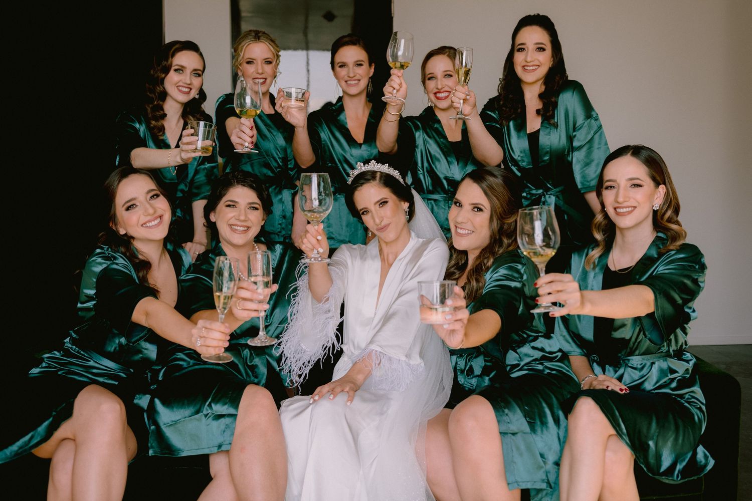 Bride in white robe with bridesmaids in green satin robes toasting champagne glasses together.
