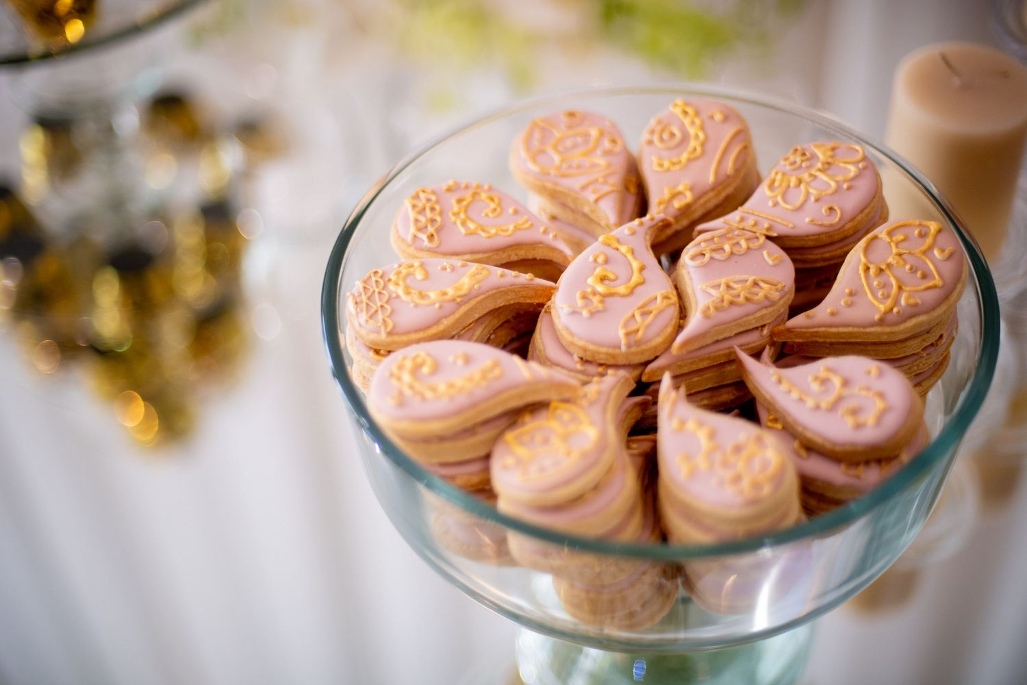 Pink heart-shaped cookies with gold floral designs displayed in a glass pedestal bowl at a wedding reception.