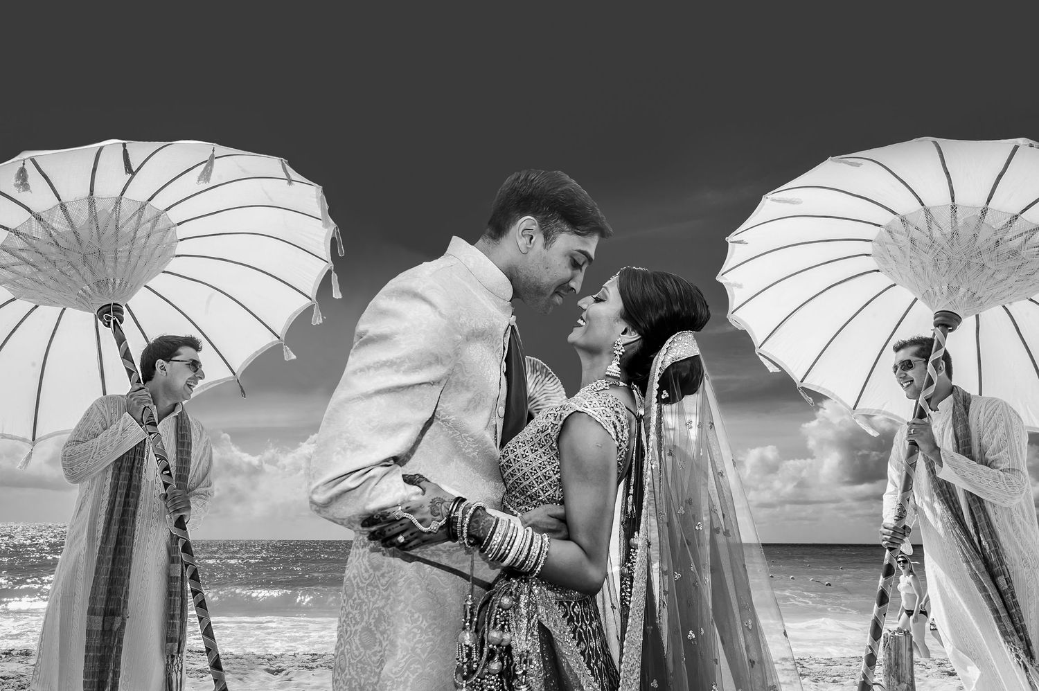 Black and white photo of Indian couple in traditional wedding attire sharing romantic moment under white umbrellas by the sea.