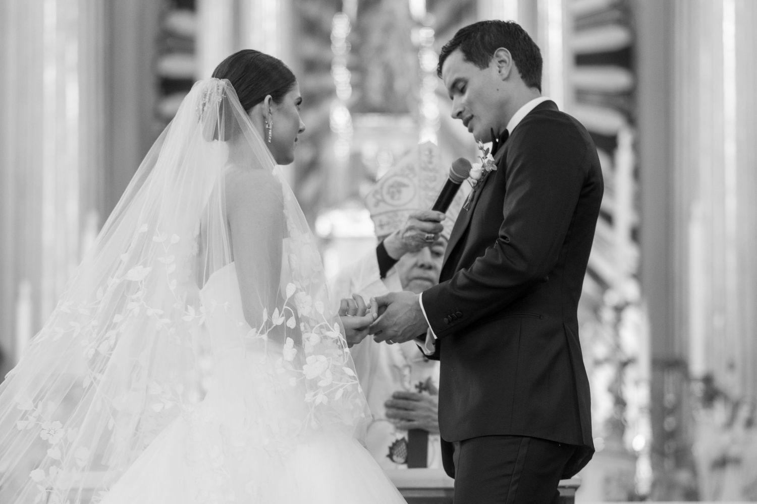 Black and white photo of a groom placing a wedding ring on his brides finger during a church ceremony.