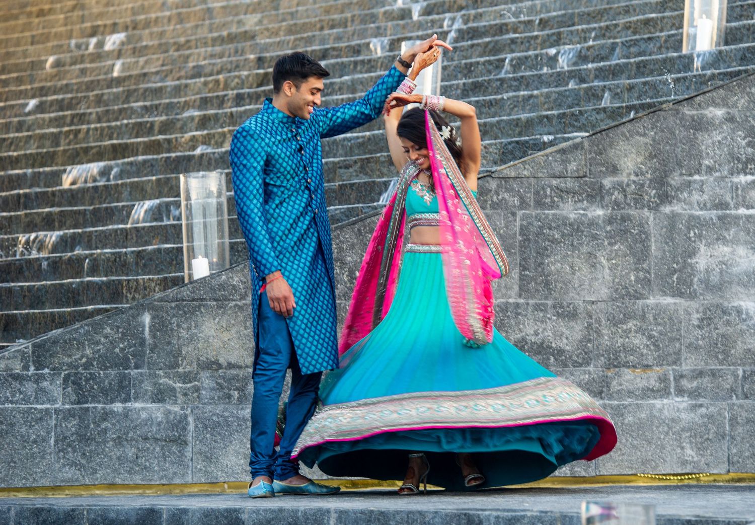 Couple in traditional Indian attire dancing on steps, woman in turquoise lehenga twirling, man in blue kurta.