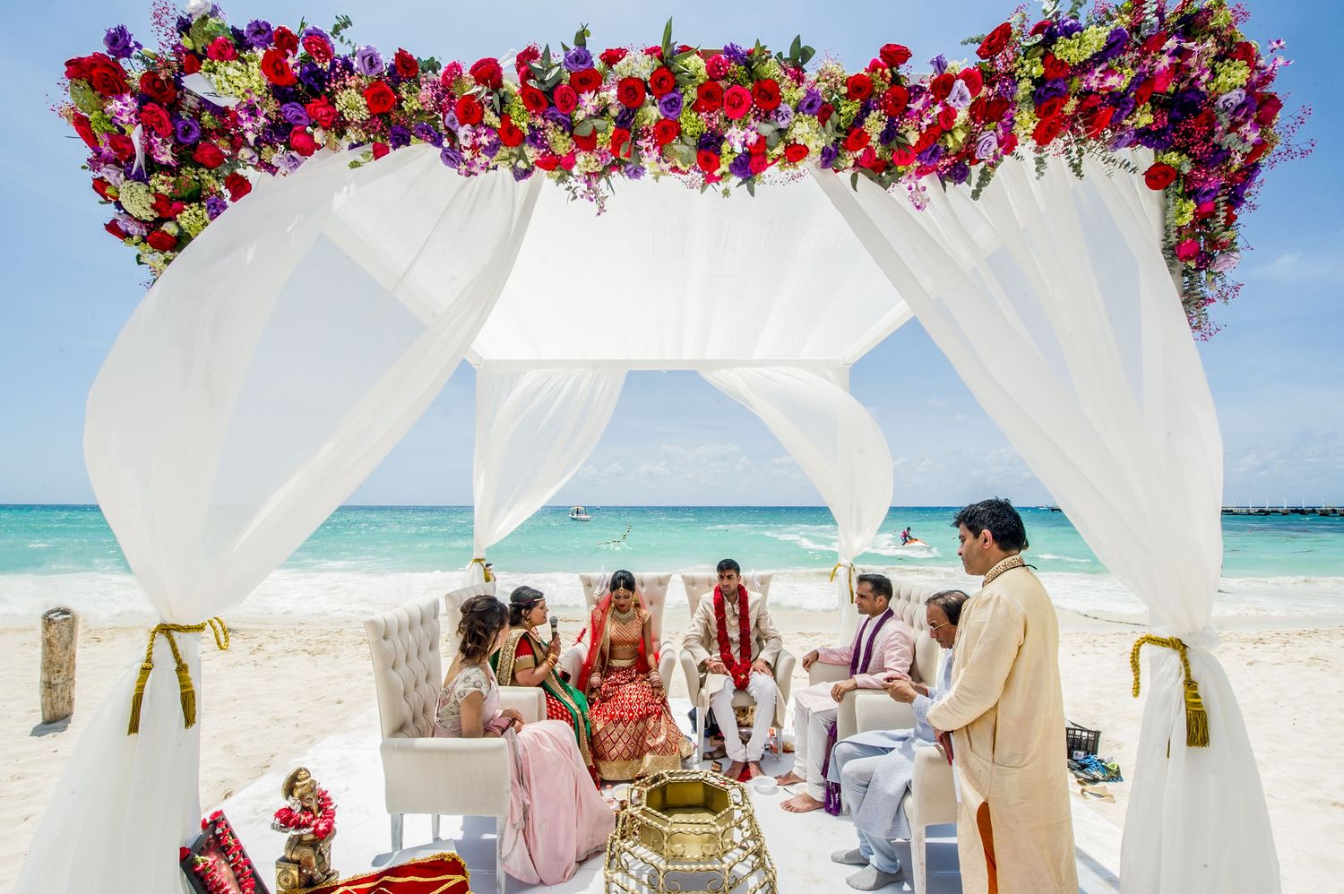 Colorful Indian beach wedding ceremony under white floral canopy with red roses on tropical white sand beach.