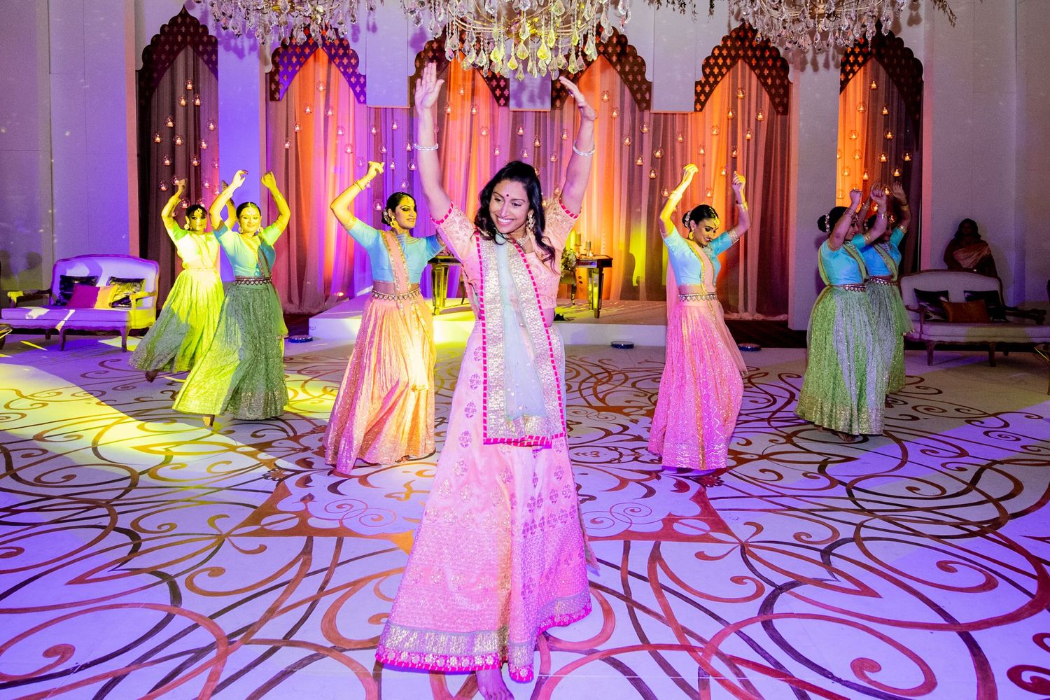 Woman in pink lehenga leads dancers in colorful traditional Indian attire at a vibrant wedding sangeet ceremony.