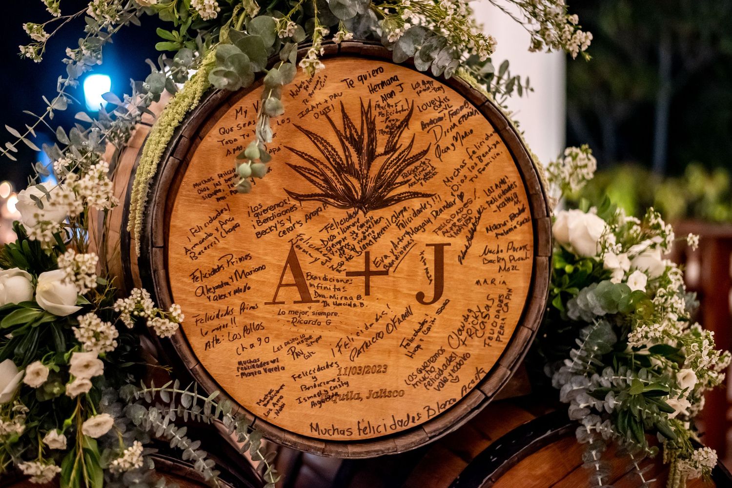 Rustic wooden barrel wedding guest book engraved with A+J initials and agave plant design, surrounded by white floral arrangements.
